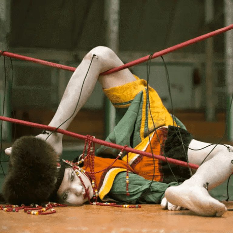 A performance photograph in which a person lies with their face on the floor, their bare legs are flung forwards over a net which they are tangled in. They are very pale, wear a fluffy brown had and a a green, yellow and orange outfit.