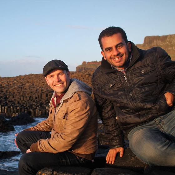 Tom and Beshoy sit on the rocks next to the Irish sea smiling towards the camera