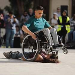 Mahmoud tilts wheel chair during a performance over a woman laying on the ground