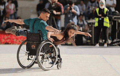 One performer with arms outstretched sits in a wheelchair. Another performer lies across him with one arm outstretched, her other hand is on the wheel.