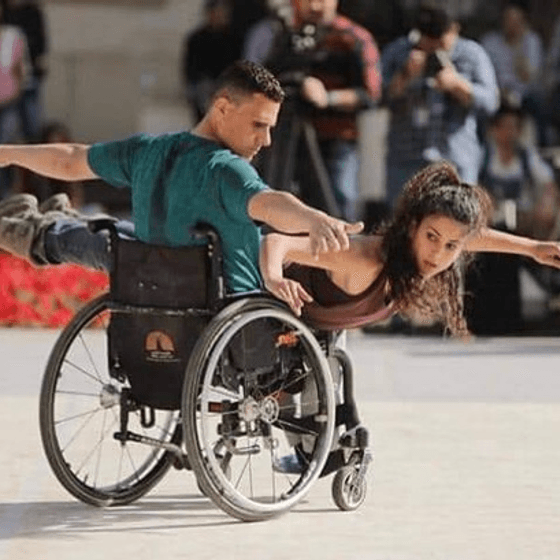 One performer with arms outstretched sits in a wheelchair. Another performer lies across him with one arm outstretched, her other hand is on the wheel.