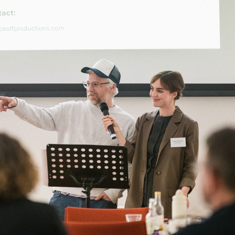 Two people, one with short brown hair and a green blazer, and the other with short white hair, a baseball cap and glasses, stand on a stage in-front of a presentation.