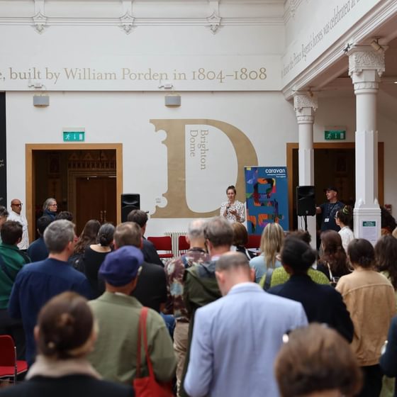 An audience stands in front of a stage where a woman holds a mic, a sign behind reads 'Brighton Dome'.