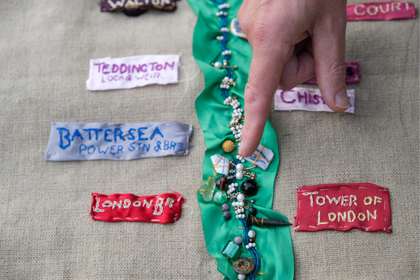 A finger points to the middle of a stitched image. A ribbon of green and beads flows down the middle of a rough canvas background, titles of Thames-side places are stitched on either side: London Bridge, Tower of London, Battersea Power Station.
