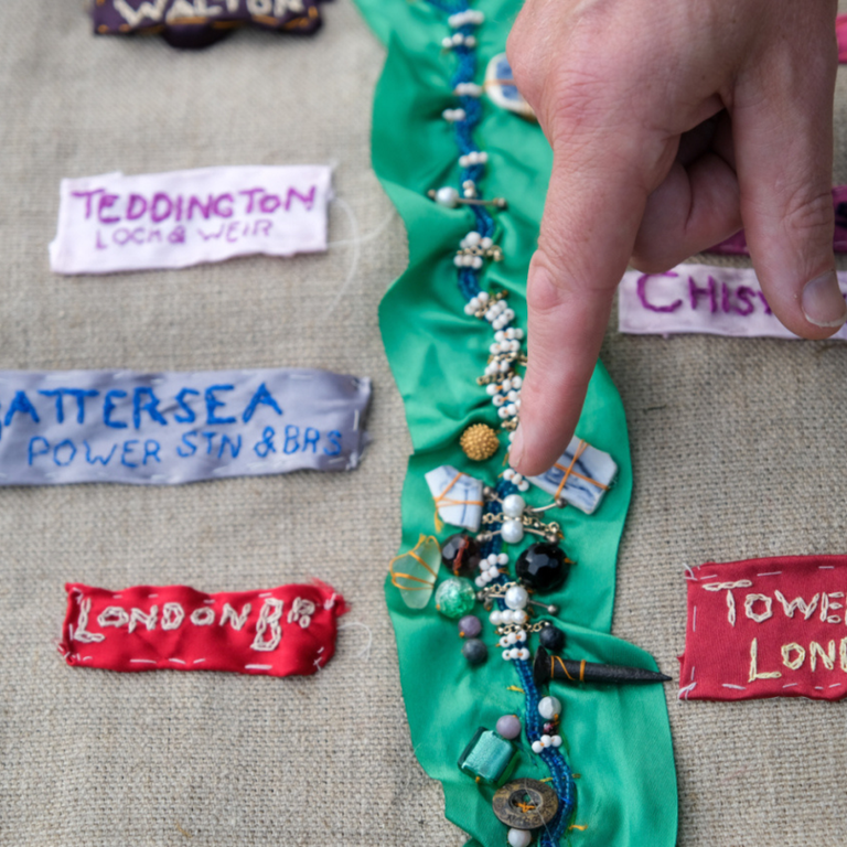 A finger points to the middle of a stitched image. A ribbon of green and beads flows down the middle of a rough canvas background, titles of Thames-side places are stitched on either side: London Bridge, Tower of London, Battersea Power Station.